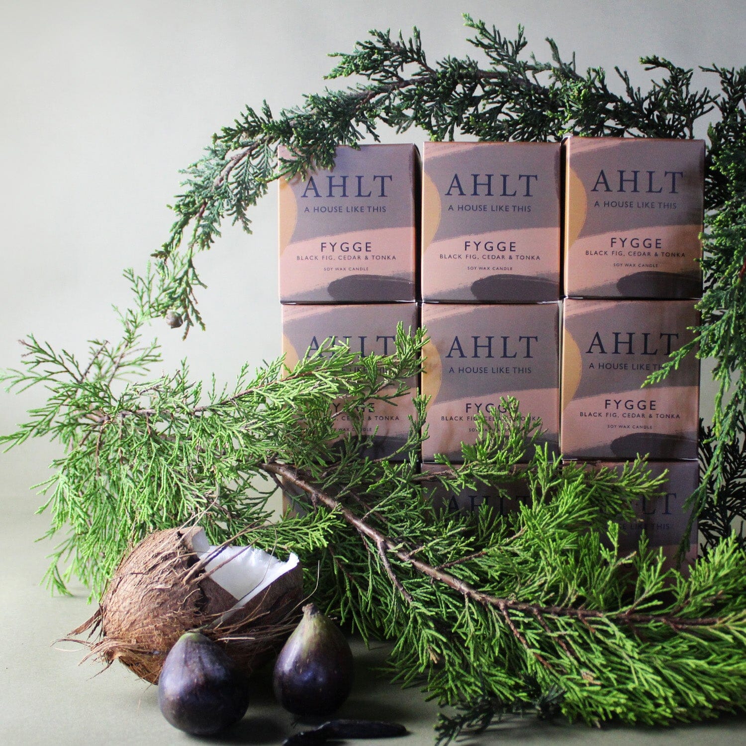 Stack of AHLT FYGGE soy candles scented with black fig, cedar, and tonka, surrounded by fresh cedar branches, whole figs, and an open coconut in a natural setting.